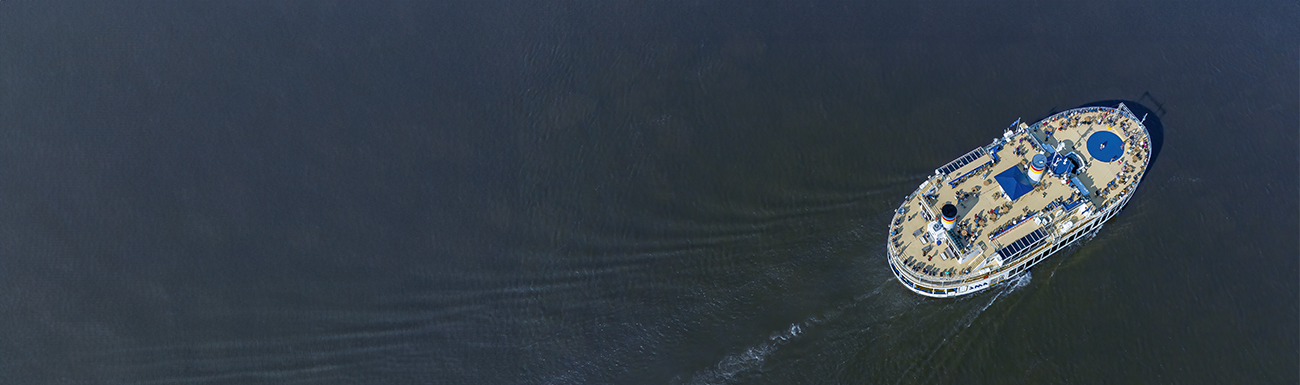 Vue aérienne sur le Louis Jolliet sur une vaste étendue d'eau bleu du St-Laurent. Le bateau avance en laissant derrière lui de légères ondulations. Il possède plusieurs ponts avec des structures visibles et des passagers à bord.